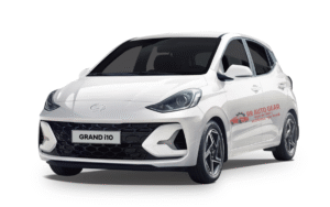 Front and side view of the 2025 Hyundai Grand i10 in red color, parked on an urban road showing its sleek design, alloy wheels, LED DRLs, and modern styling features.