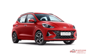 Front and side view of the 2025 Hyundai Grand i10 in red color, parked on an urban road showing its sleek design, alloy wheels, LED DRLs, and modern styling features.
